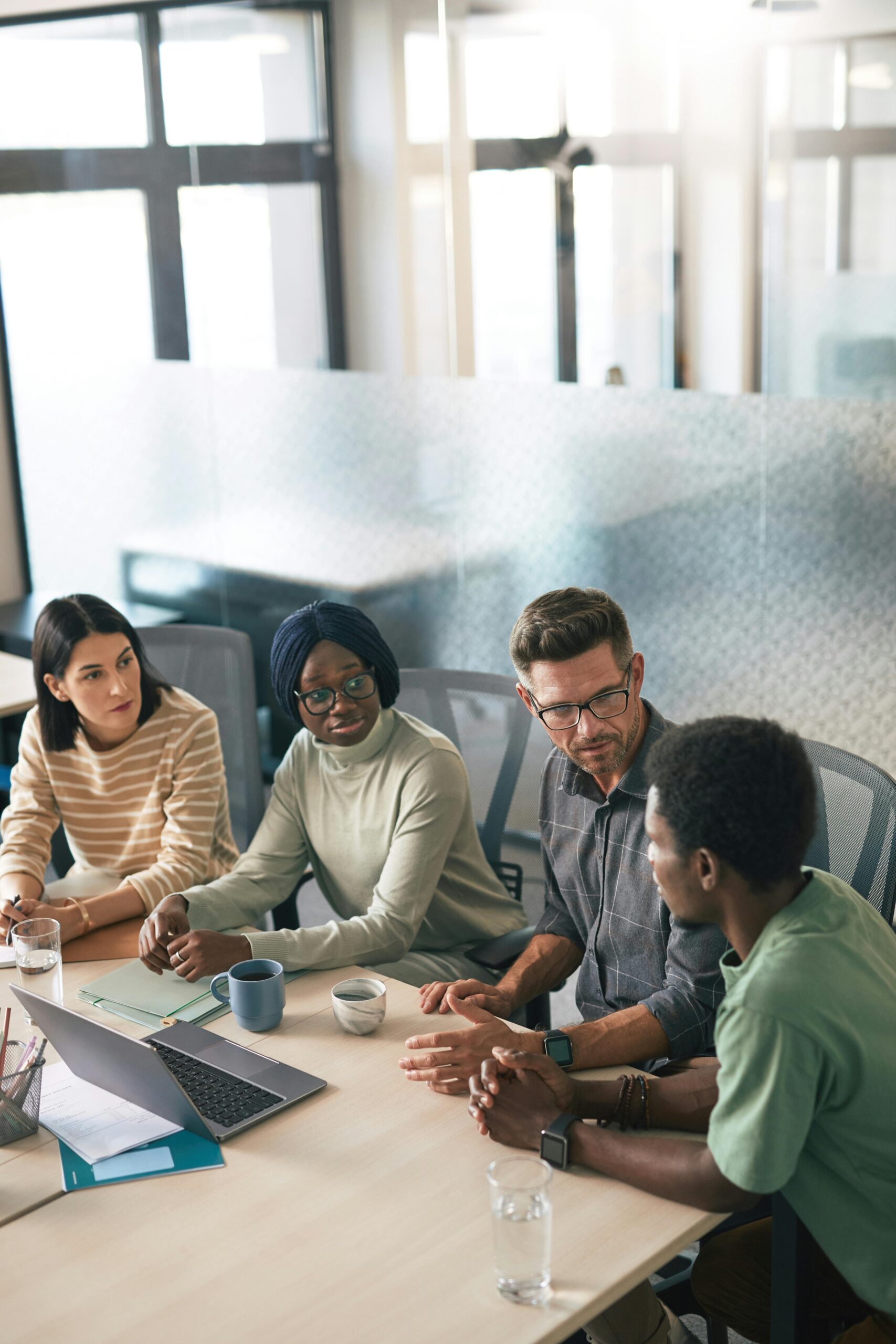 A diverse group of colleagues having a discussion in a modern office setting with laptops and coffee cups.